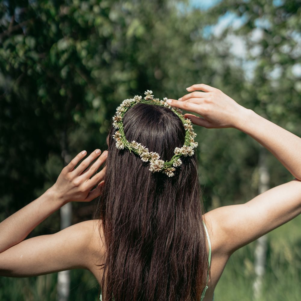 Back view of woman with flower crown in nature, hands on head, wearing a floral dress.