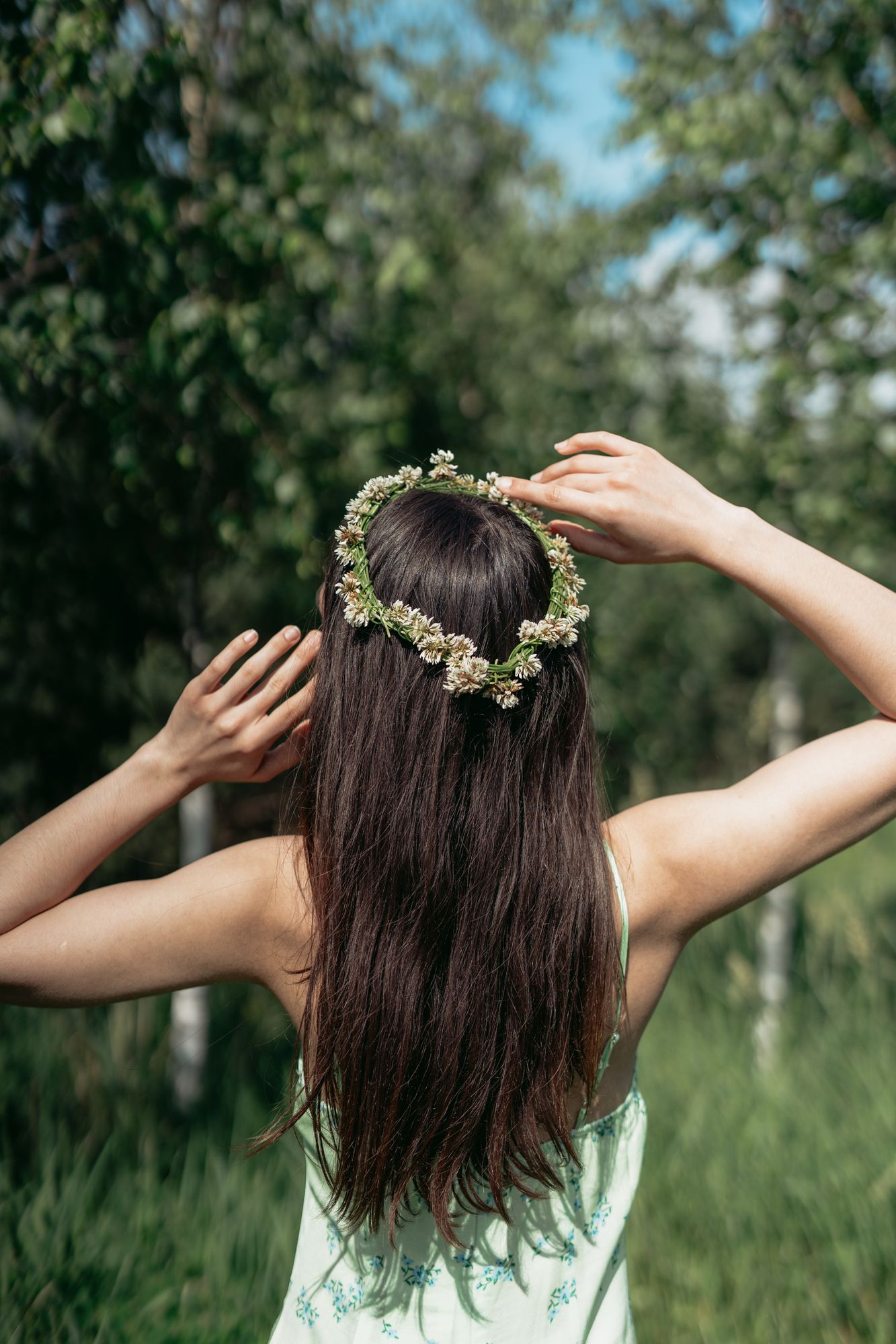 Back view of woman with flower crown in nature, hands on head, wearing a floral dress.